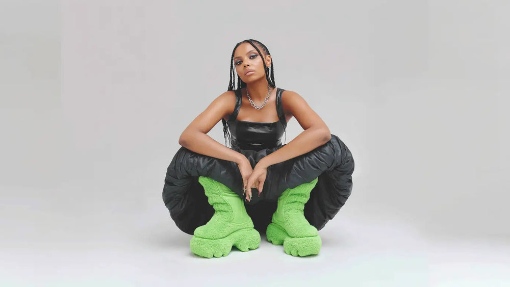 Confident woman in a high-fashion studio portrait, crouching against a light gray background, wearing a black leather top and voluminous black pants with bold neon green fuzzy platform boots.